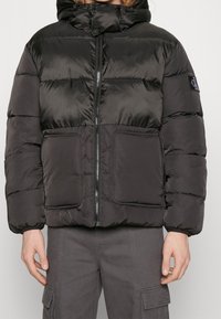 Czarna kurtka puchowa z kapturem, zapinana na zamek z przodu, z dużymi naszywanymi kieszeniami oraz błyszczącym materiałem z pikowanym wzorem.