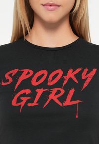 Acercamiento a una mujer vestida con una camiseta negra con el texto rojo en negrita "SPOOKY GIRL" en una fuente desgastada y goteante.