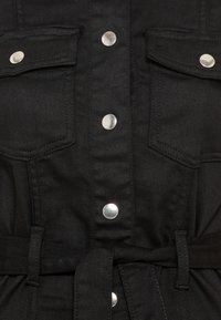 Svart denimjacka med silverknappar, två bröstfickor, bälte i midjan och slät textur. Fokusera på funktionella designelement.