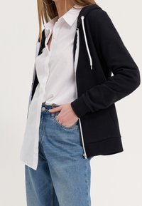 Personne portant une chemise blanche boutonnée, un sweat à capuche noir zippé avec des cordons blancs, et un jean bleu, avec une main dans la poche.