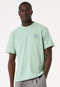 Lichtgroene katoenen t-shirt met een ronde hals, korte mouwen en een blauw logo op de linkerborst. Zachte textuur, casual pasvorm.