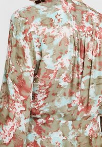 Blouse à motifs floraux en tissu doux, présentant des couleurs pastel de vert, rouge et bleu. Le design comprend un pli dans le dos et une ceinture.