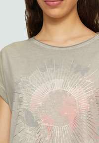 Camiseta de manga corta color gris claro con escote redondo. Presenta un diseño circular central en tonos de rosa y plata, con sutiles detalles de mariposas.