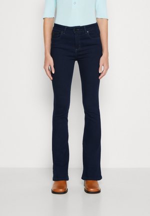 Mujer con vaqueros bootcut azul oscuro, top azul claro de manga tres cuartos y botines de cuero marrón, de pie sobre fondo blanco.