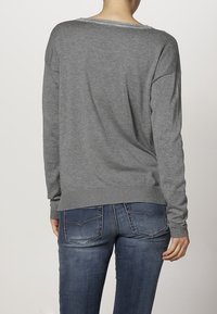 Grauer Pullover mit langen Ärmeln, aus weichem Stoff; hat einen runden Ausschnitt und einen gerippten Saum. Kombiniert mit blauen Jeans.
