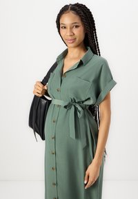 Abito verde con bottoni e maniche corte, dotato di una cintura in vita e due tasche sul petto. Il modello porta una borsa a tracolla nera.