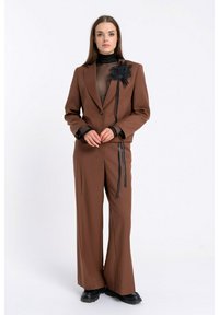 Brązowy garnitur z szerokimi spodniami i dopasowaną marynarką, z czarnym kwiatowym akcentem na klapie oraz przezroczystą czarną bluzką pod spodem.