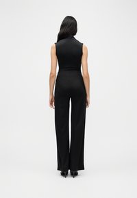 Γυναίκα με μακριά σκοτεινά μαλλιά που φοράει μαύρο αμάνικο τοπ, μαύρο παντελόνι με ψηλή μέση και φαρδιά μπάρα, και μαύρες γόβες, με την πλάτη προς τα πίσω σε λευκό φόντο.