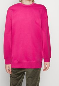 Fuchsia sweatshirt med långa ärmar, ribbade manschetter och rundad hals. Gjord av mjukt tyg med en slät textur. Kombinerad med olivgröna byxor.