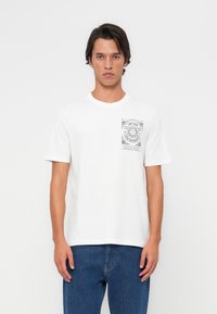 T-shirt en coton blanc avec un motif imprimé sur la partie gauche de la poitrine. Manches courtes et col rond.