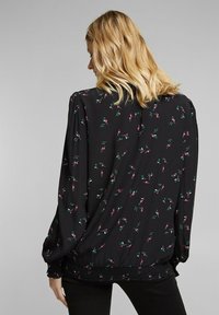 Schwarze Bluse mit langen Ärmeln und gerafftem Saum, die kleine rosa und grüne Blumenmuster auf einem glatten Stoff aufweist.