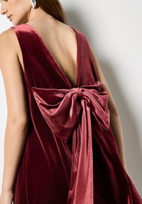 Bordowa aksamitna sukienka z głębokim dekoltem na plecach, ozdobiona dużą kokardą z tyłu. Gładka tekstura z błyszczącymi akcentami na powierzchni.