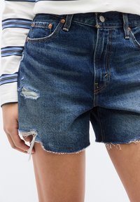 Κοντινή λήψη σκισμένου μπλε τζιν σορτς που φοριέται με μακρυμάνικη μπλούζα λευκή με μπλε ρίγες, εμφανίζοντας ξεφτισμένες άκρες και το χέρι τοποθετημένο στο πλάι.