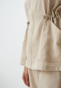 Beige Jacke mit Reißverschluss, gerafften Seiten und großen Taschen. Hergestellt aus glattem Stoff mit lockerer Passform und subtiler Textur.