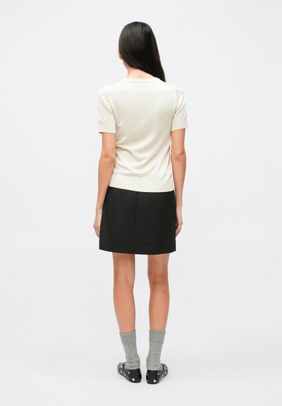 Haut ajusté beige clair à manches courtes, associé à une jupe noire. Des chaussettes grises en maille et des chaussures noires avec des accents métalliques sont visibles.