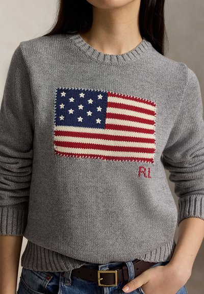 Personne portant un pull gris avec un motif tricoté du drapeau américain sur la poitrine et un jean bleu avec une ceinture marron.
