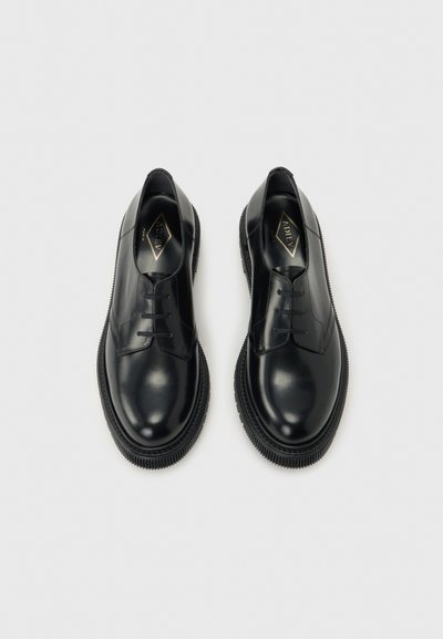 Paire de chaussures habillées noires en cuir brillant à lacets, avec semelles épaisses rainurées, vues de dessus sur un fond gris clair.