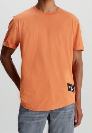 Homme portant un t-shirt orange à manches courtes avec des poignets retroussés et un petit patch logo Calvin Klein noir et blanc près de l'ourlet, associé à un jean gris.