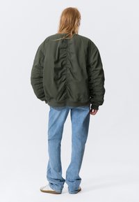 Personne avec de longs cheveux rouge vif portant une grande veste verte foncée, un jean bleu clair et des baskets blanches, se tenant de dos sur un fond blanc.