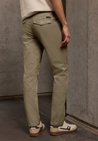 Homme portant un pantalon beige et des baskets blanches, debout avec la main droite détendue le long du corps, appuyé contre un mur en béton texturé.