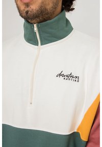 Maglione con cerniera a un quarto in colori crema, verde, senape e granata. Caratterizzato da logo ricamato e tessuto morbido con una texture liscia.