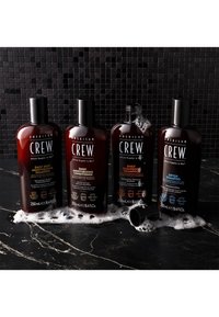 Neljä ruskeaa pulloa American Crew -hiustuotteita mustilla kansilla ja etiketeillä, joilla on erilaisia shampoo- ja hoitoainetyyppejä. Kuplat ympäröivät niitä.
