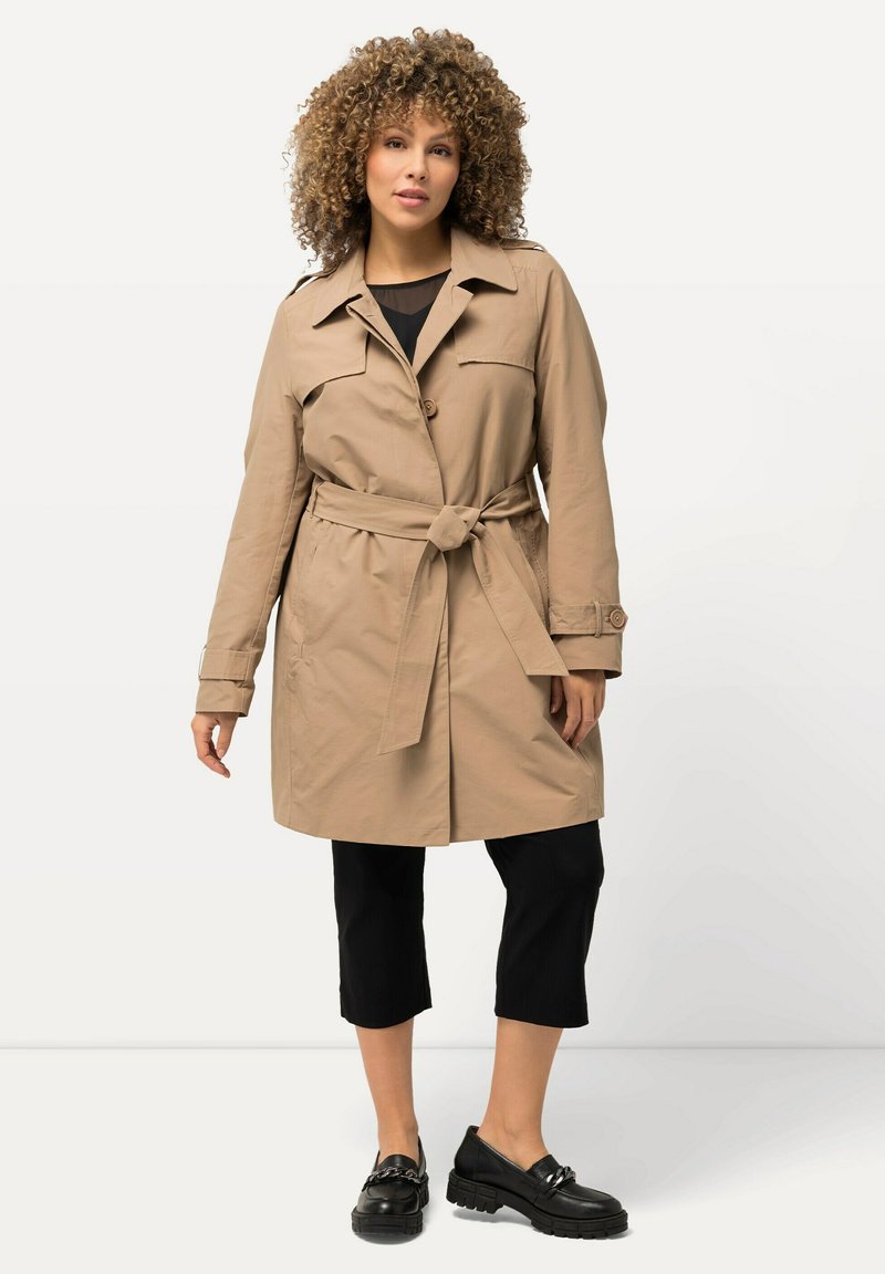 Trench beige con cinturón en la cintura, gran cuello y cierre de botones. Usado sobre pantalones negros recortados y zapatos negros de suela gruesa.