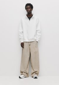 Witte kwart-rits sweatshirt, relaxed fit, gecombineerd met oversized beige jeans. Model draagt witte en zwarte sneakers. Eenvoudig, strak design.