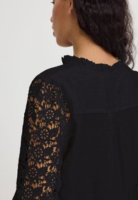 Blusa nera con intricate maniche lunghe in pizzo nero, scollo a volant e corpo solido con una texture liscia, vista dal retro.