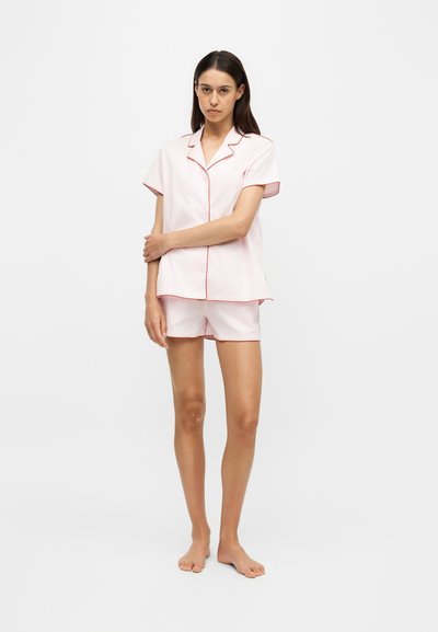 Jeune femme debout pieds nus portant un haut de pyjama rose clair à manches courtes et un short assorti avec une bordure rouge sur un fond blanc uni.