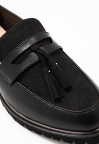 Mocassin noir en matériaux en daim et cuir, avec un accent de pompon, une forme arrondie et une semelle extérieure en caoutchouc texturé.