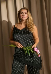 Γυναίκα με μακριά ξανθά μαλλιά, φορώντας μαύρο σατέν τοπ και παντελόνι με πουά, κρατά τρία ροζ άνθη παιώνιας, στέκεται μπροστά από μπεζ κουρτίνες.