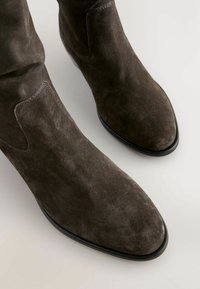 Bottines en daim marron avec un bout pointu, des panneaux latéraux élastiques et une semelle lisse noire, présentant une texture douce et des lignes épurées.