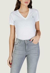 Femme portant un t-shirt blanc à col en V et manches courtes avec une broche argentée en forme de "A" et un jean taille haute gris clair, main dans la poche.
