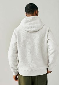 Sudadera blanca con patrón floral texturizado, bolsillo tipo canguro y puños acanalados. Presenta una capucha espaciosa y una tela suave.