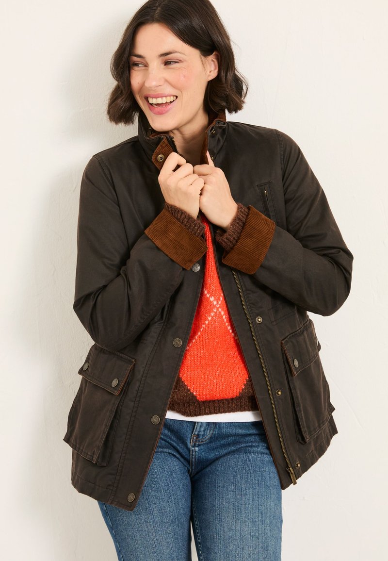 Woman smiling, holding collar of dark brown jacket with corduroy cuffs, wearing blue jeans and orange patterned sweater underneath.