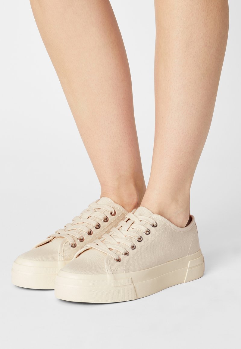 Pieds portant des baskets basses en toile beige avec des semelles épaisses en caoutchouc et des lacets, debout sur un fond blanc.