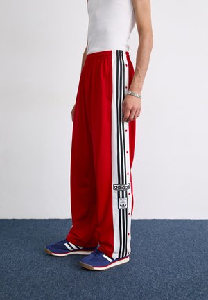 Røde sportbukser med sorte og hvide side-striber, knaplukninger og oversized pasform. Båret med blå sneakers med hvide striber.