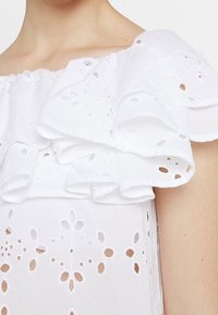 Vit blus med volangdetalj över axlarna, broderade ögonluckor och blomformade utskärningar genom tyget, med en mjuk textur.