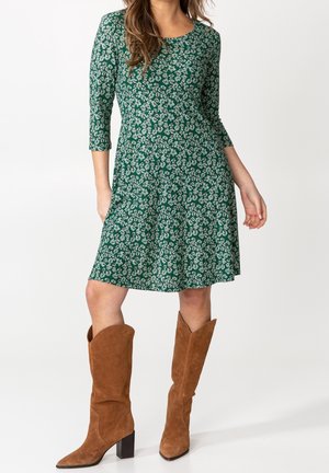 Vrouw draagt een groene bloemenprint jurk tot op de knie met driekwart mouwen en bruine suède mid-kuitlaarzen met hak.