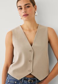 Beige ärmellose Weste mit V-Ausschnitt, ausgestattet mit drei Knöpfen an der Vorderseite, einer taillierten Passform und weichem Stoff, kombiniert mit hochgeschnittenen Jeans.