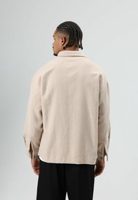 Beige oversized overhemd met kraag, gestructureerde stof en zijknopaccenten, gezien van achteren, gecombineerd met donkere broek.