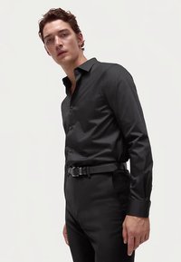 Zwart, nauwsluitend shirt met lange mouwen en een kraag met knoopjes, gladde textuur en contrasterende knopen. Gecombineerd met op maat gemaakte zwarte broek.