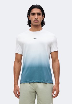 Jeune homme aux cheveux foncés mi-longs portant un t-shirt Reebok dégradé blanc-bleu et un short beige clair, debout, faisant face à l’avant.