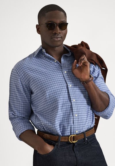 Niebieska koszula w kratę na guziki z podwiniętymi rękawami, brązowy pasek ze złotą klamrą i ciemne dżinsy. Okulary przeciwsłoneczne na nosie, brązowa kurtka zarzucona na ramię.