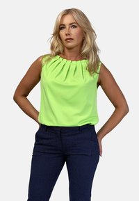 Neon zielona bluzka bez rękawów z plisowanym dekoltem, w zestawieniu z granatowymi spodniami w kropki. Gładki materiał i dopasowany krój.