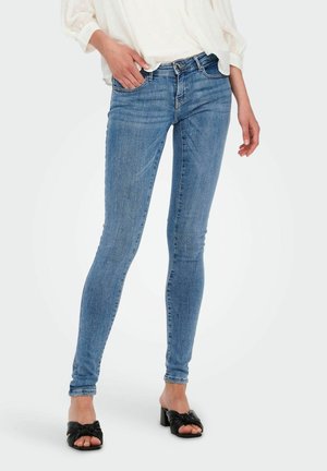 Kvinde iført lyseblå skinny jeans, hvid bluse og sorte åbne hælede sandaler står med den ene hånd i lommen.