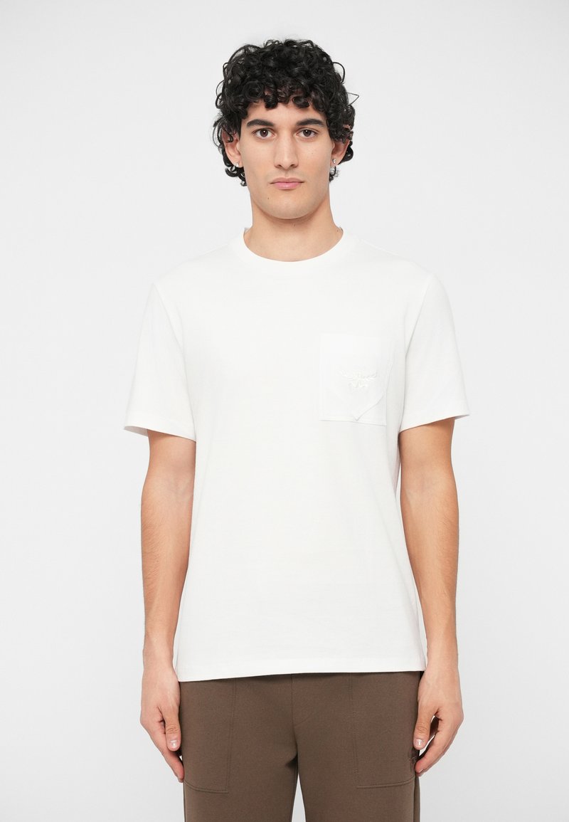 Young man with curly dark hair wearing a plain white short-sleeve T-shirt with a chest pocket and brown pants, standing against white background.