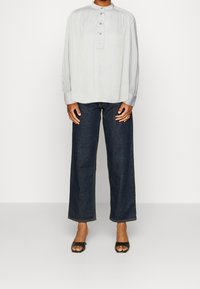 Maglietta a maniche lunghe grigio chiaro con colletto alto e tre bottoni, abbinata a jeans a gamba larga blu scuro e sandali neri open-toe.