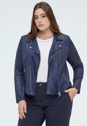 Pitkähiuksinen nainen pukeutuneena laivastonsiniseen nahkatakkiin valkoisen paidan ja tummien housujen yllä, poseeraa yhdellä kädellä taskussa.